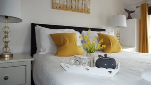 Una cama bien hecha con almohadas blancas y mostaza, una mesita de noche con lámpara, un plato decorativo y un cuadro colgado en la pared.