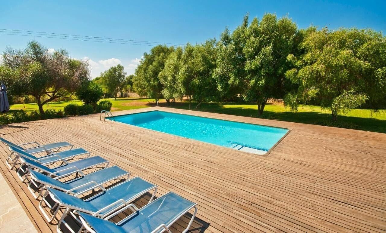 A rectangular swimming pool with a wooden deck, surrounded by trees and a row of blue lounge chairs.