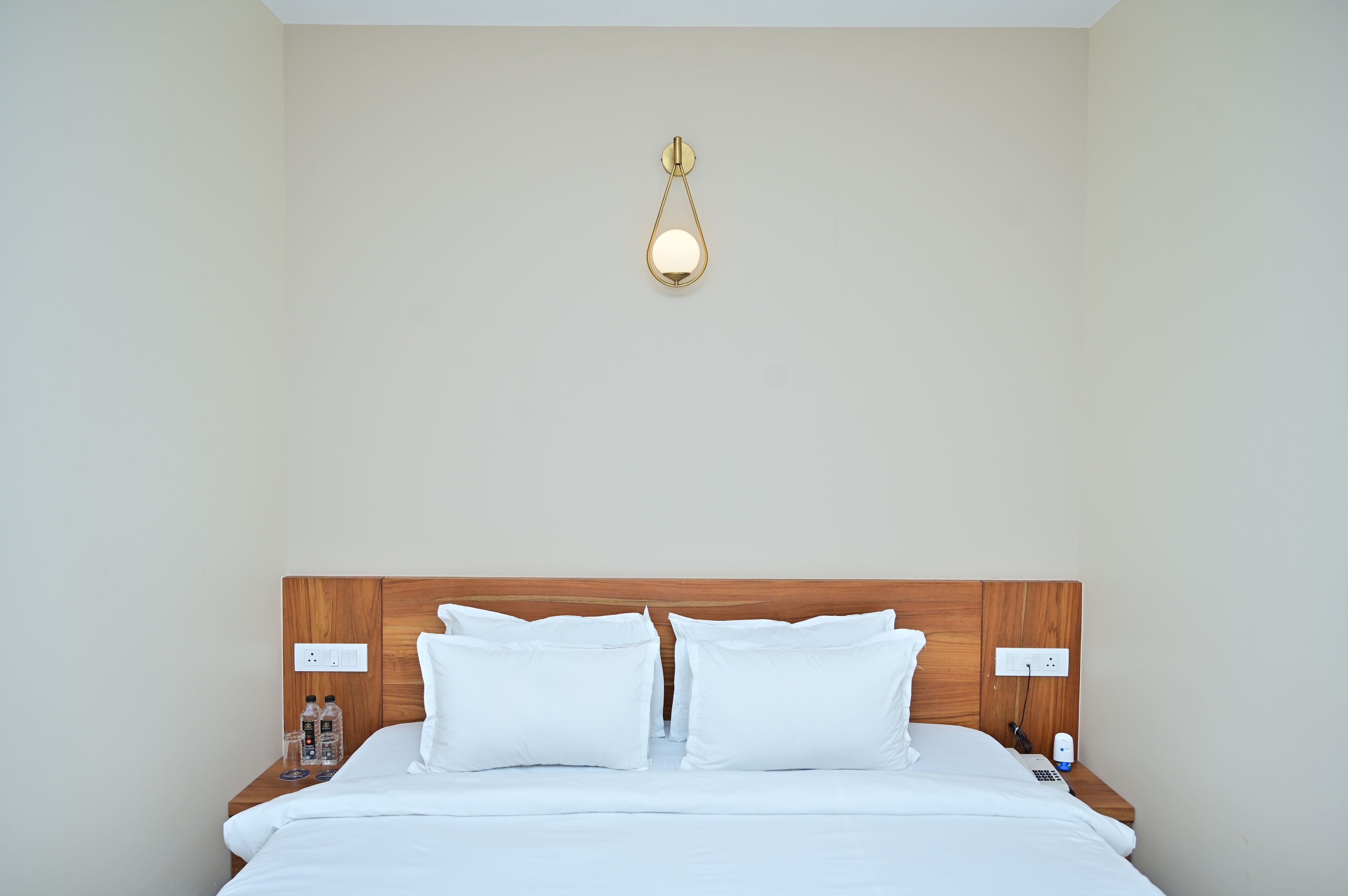 A hotel room with a wooden headboard, white bedding, a bedside table with a bottle and glasses, and a wall-mounted light fixture.