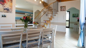 Un coin repas avec une table en bois, des chaises blanches et un vase de fleurs. Un escalier avec des rampes en bois mène à une autre pièce.