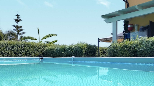 Une piscine d’un bleu éclatant, bordée de carreaux, entourée d’une végétation luxuriante et adossée à un bâtiment doté d’une zone abritée.