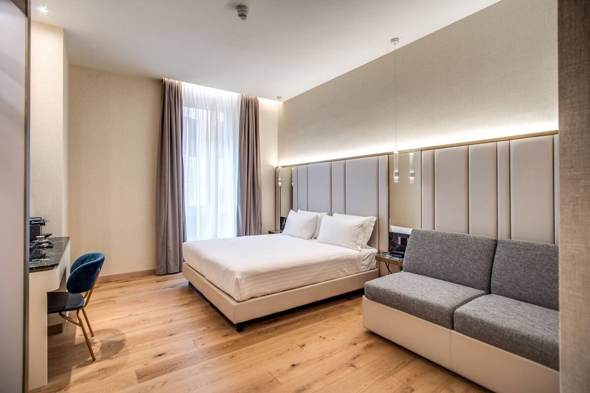 A modern hotel room with a large bed, a sofa, a desk with a chair, and a large window with curtains.