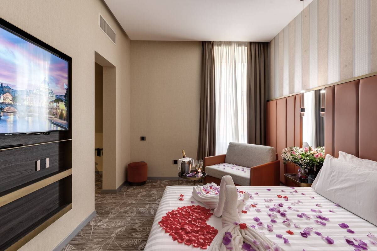 A hotel room with a bed, a TV, a chair, a small table with a bottle and glasses, and a window with curtains.