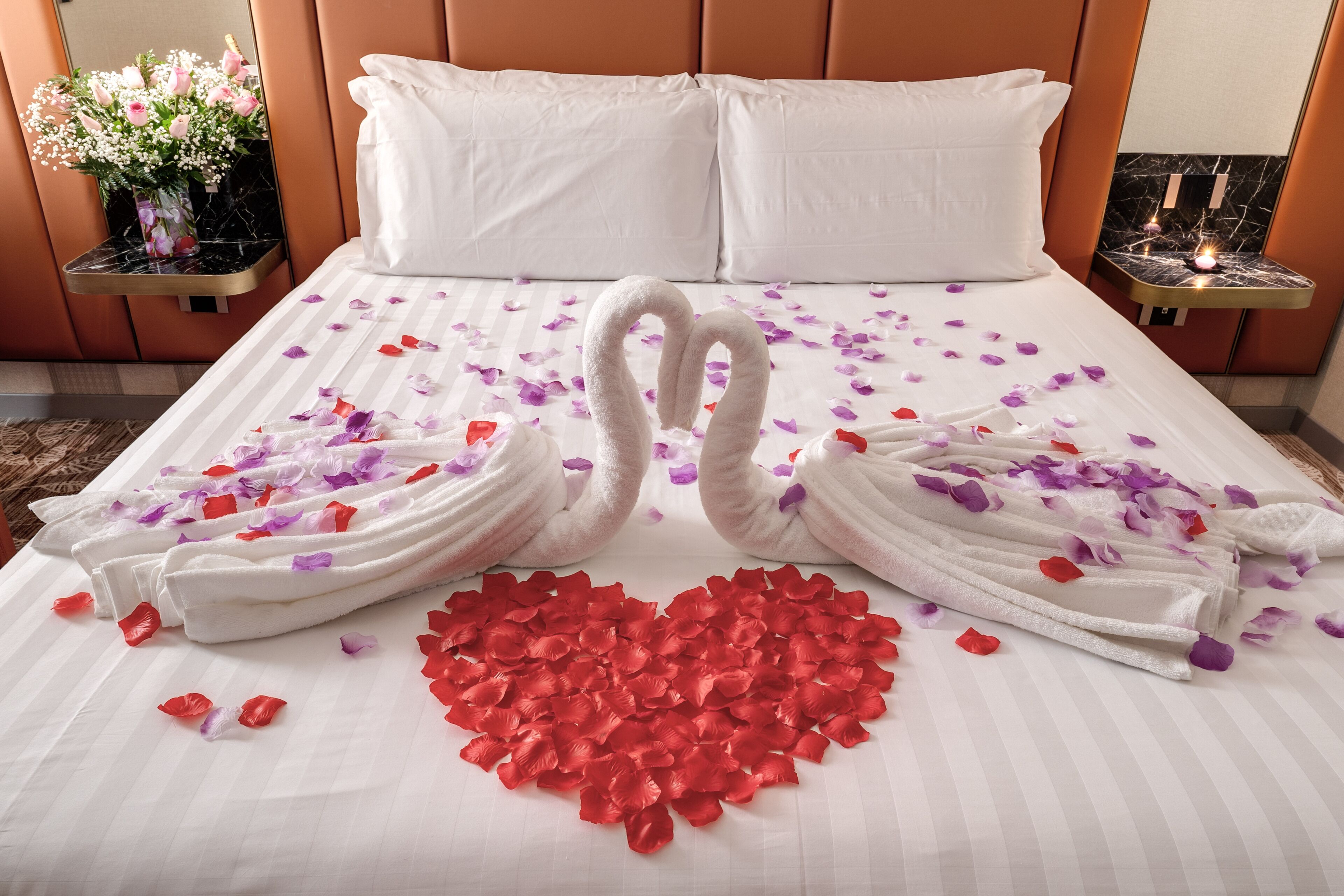 A neatly made bed with white linens, two towels arranged in a heart shape, and a heart of red rose petals.