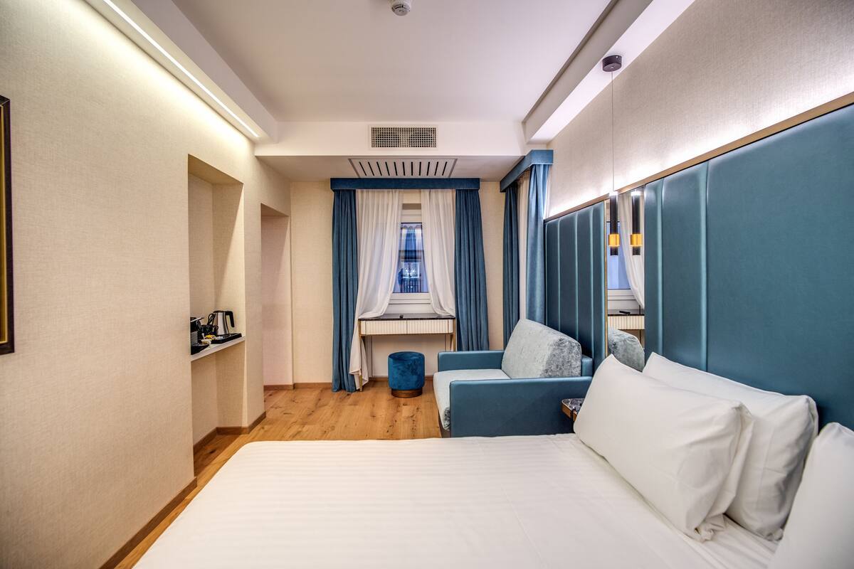 A hotel room with a large bed, a desk with a computer, a small ottoman, and a view of a building through a window with blue curtains.