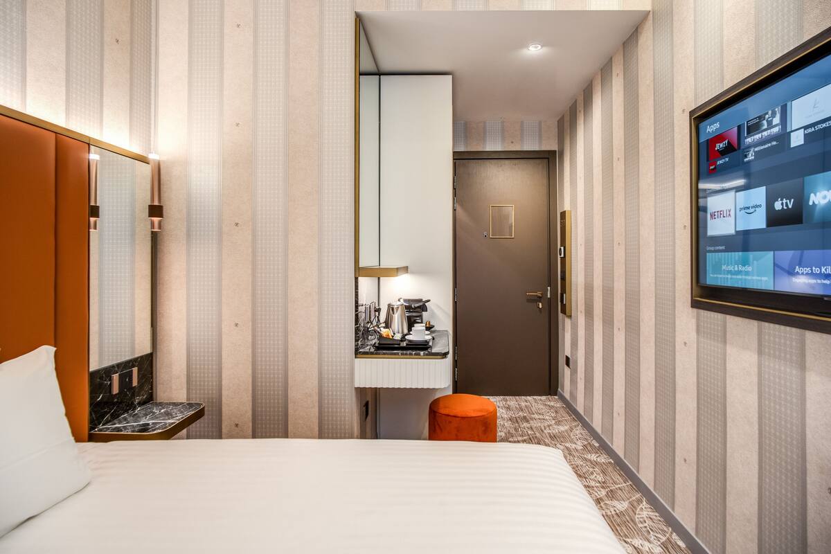 A hotel room with a bed, a bedside table, a mirror, a TV mounted on the wall, and a door leading to a bathroom.