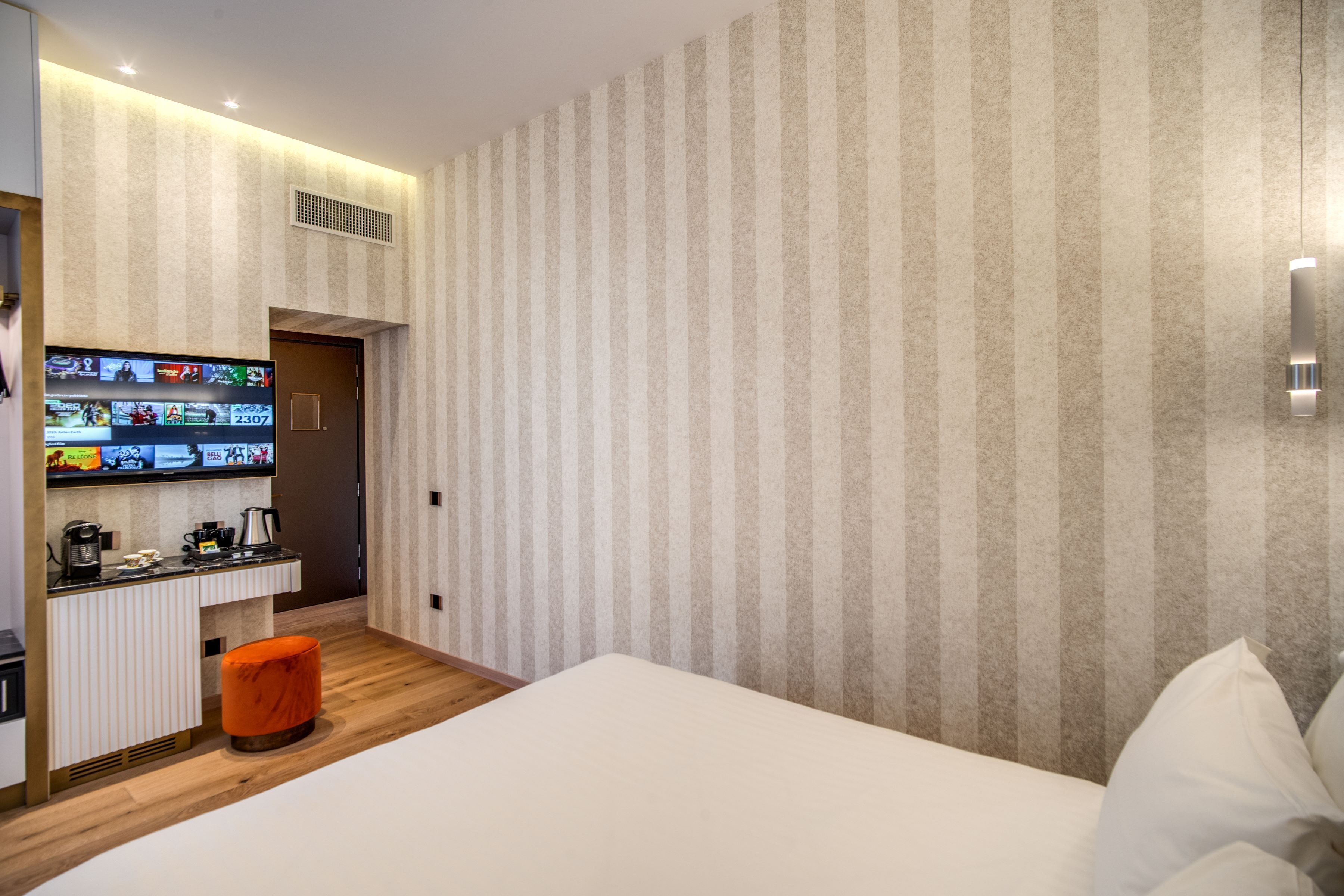 Une chambre d’hôtel moderne avec un grand lit, un petit pouf orange, un téléviseur fixé au mur et un papier peint à rayures.