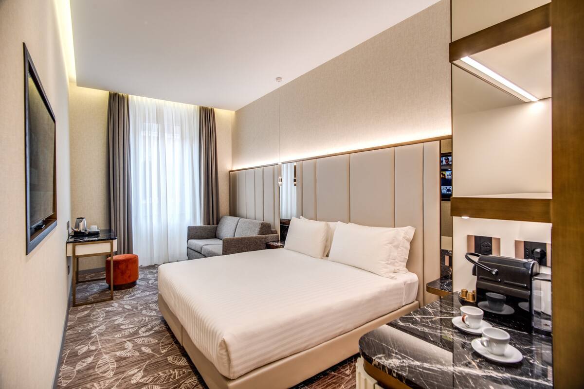 A hotel room with a large bed, a desk with a chair, a sofa, a coffee machine, and a TV.