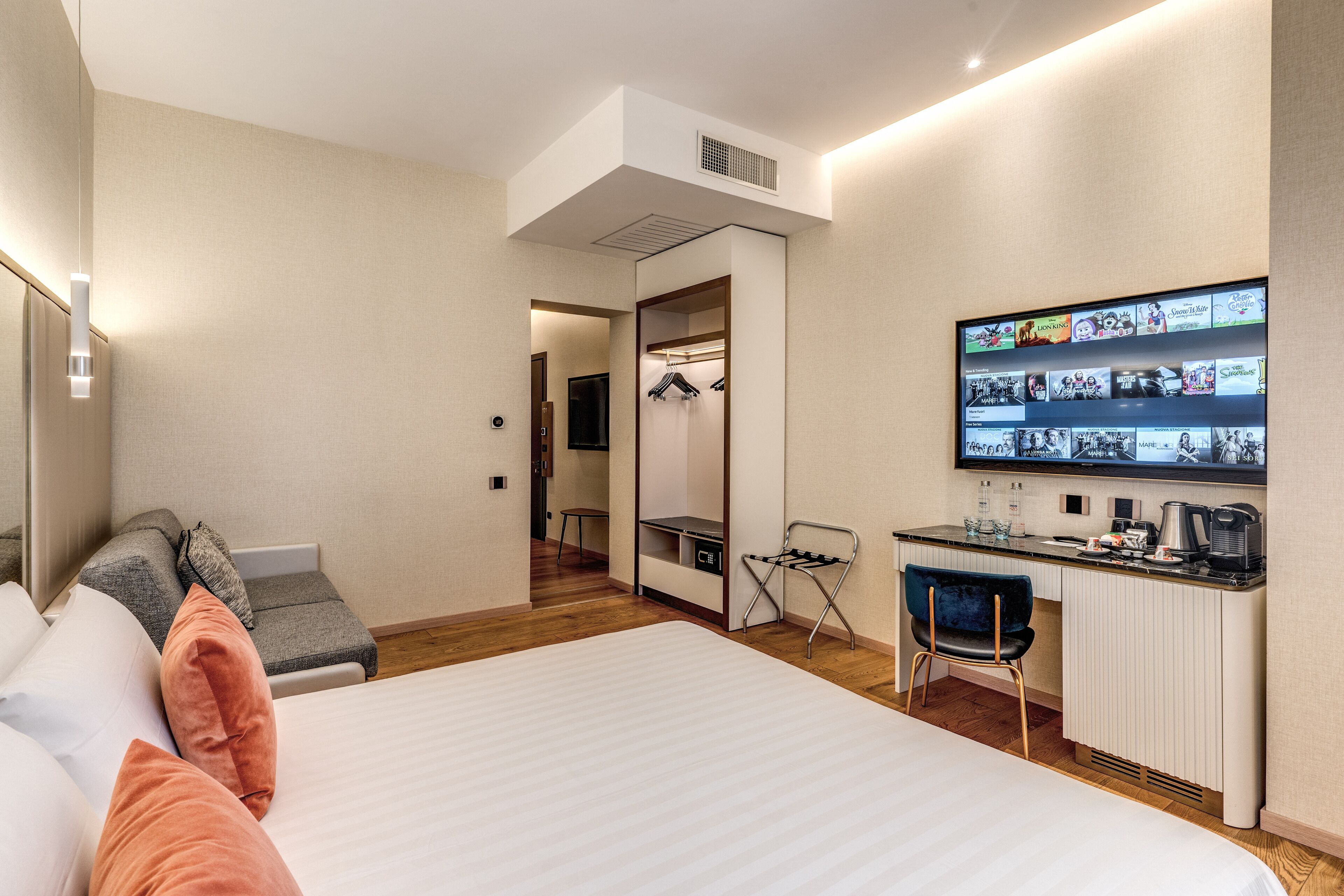 A modern hotel room with a bed, a seating area with cushions, a small desk with a chair, a mirror, and a television displaying a movie selection.
