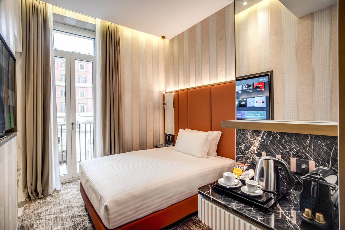 A hotel room with a bed, a TV mounted on the wall, a coffee maker, and a view of the city through the window.