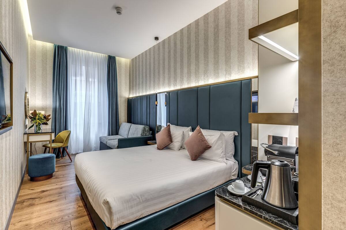 A modern hotel room with a large bed, a desk with a chair, a coffee maker, and a mirror.