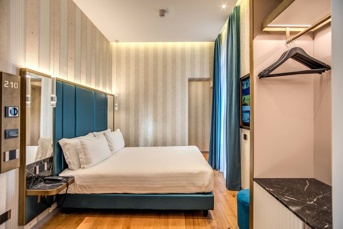 A modern hotel room with a large bed, a vanity with a mirror, a small table, and a hanging coat rack.