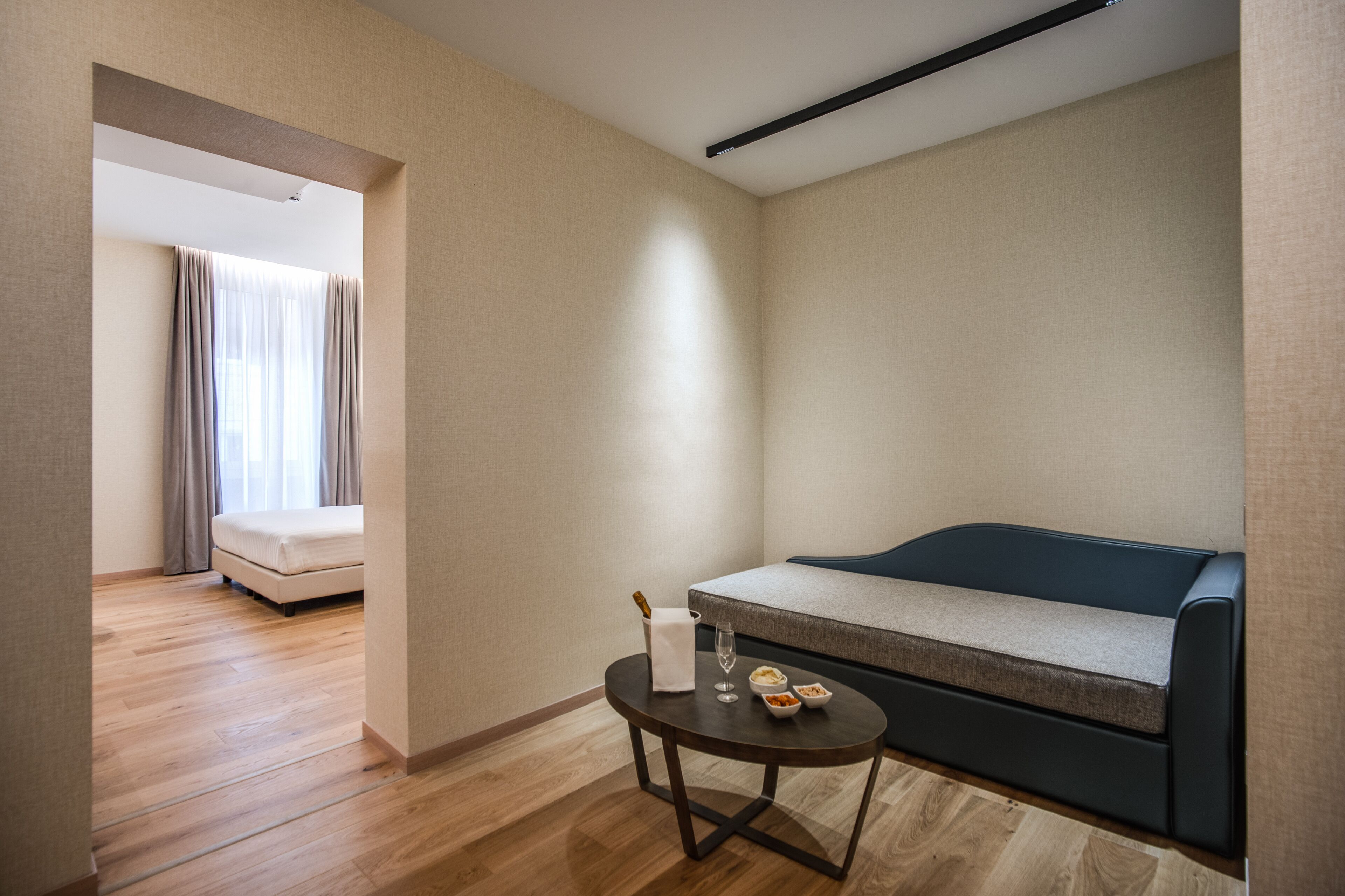 Una habitación de hotel moderna con sofá, una mesita redonda con una botella y vasos, y vistas a un dormitorio con cama y cortinas.