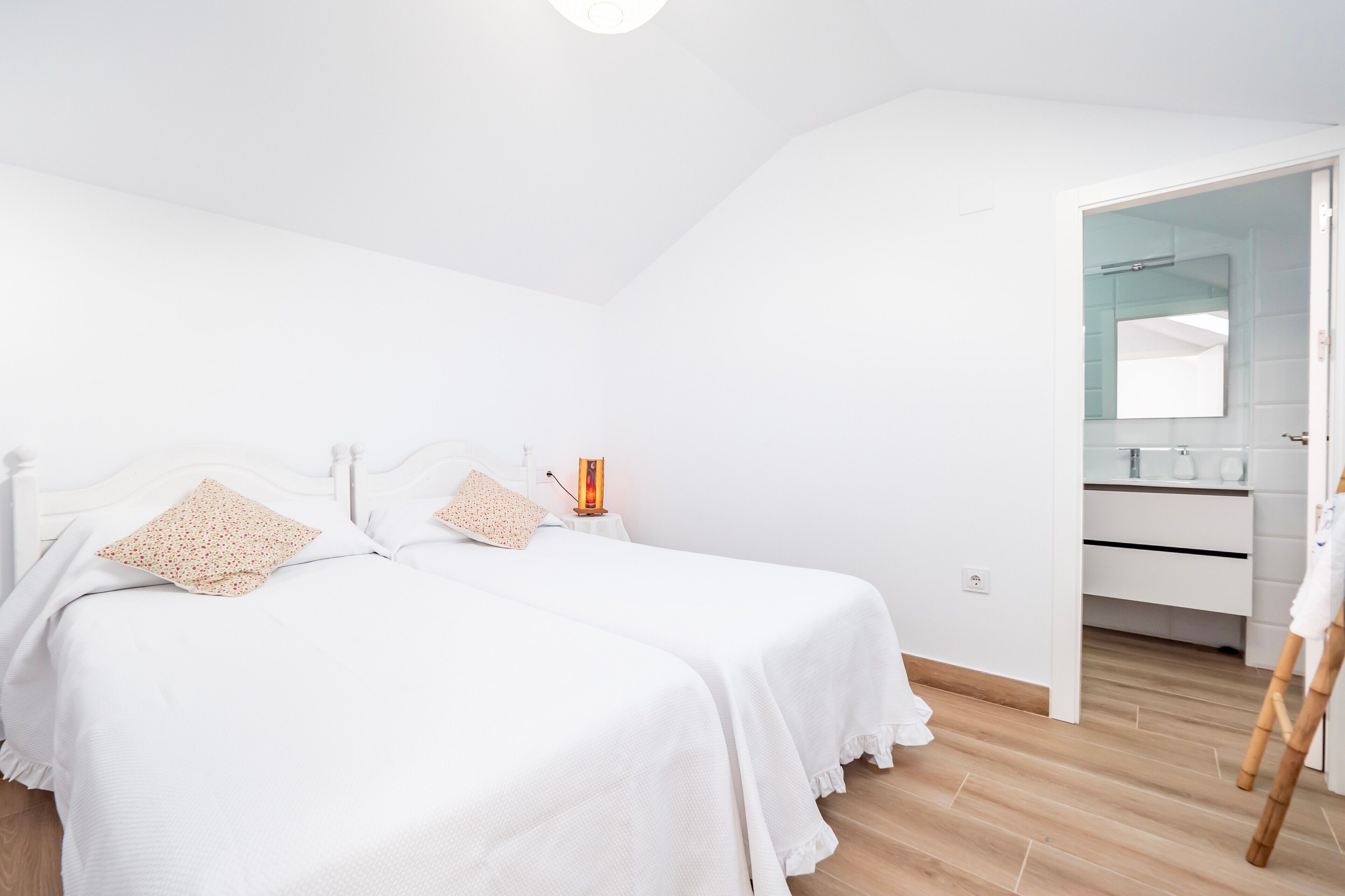 A bedroom with two beds, a bedside table with a lamp, a wooden floor, and a bathroom visible through an open door.