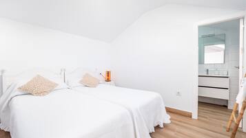 A bedroom with two beds, a bedside table with a lamp, a wooden floor, and a bathroom visible through an open door.