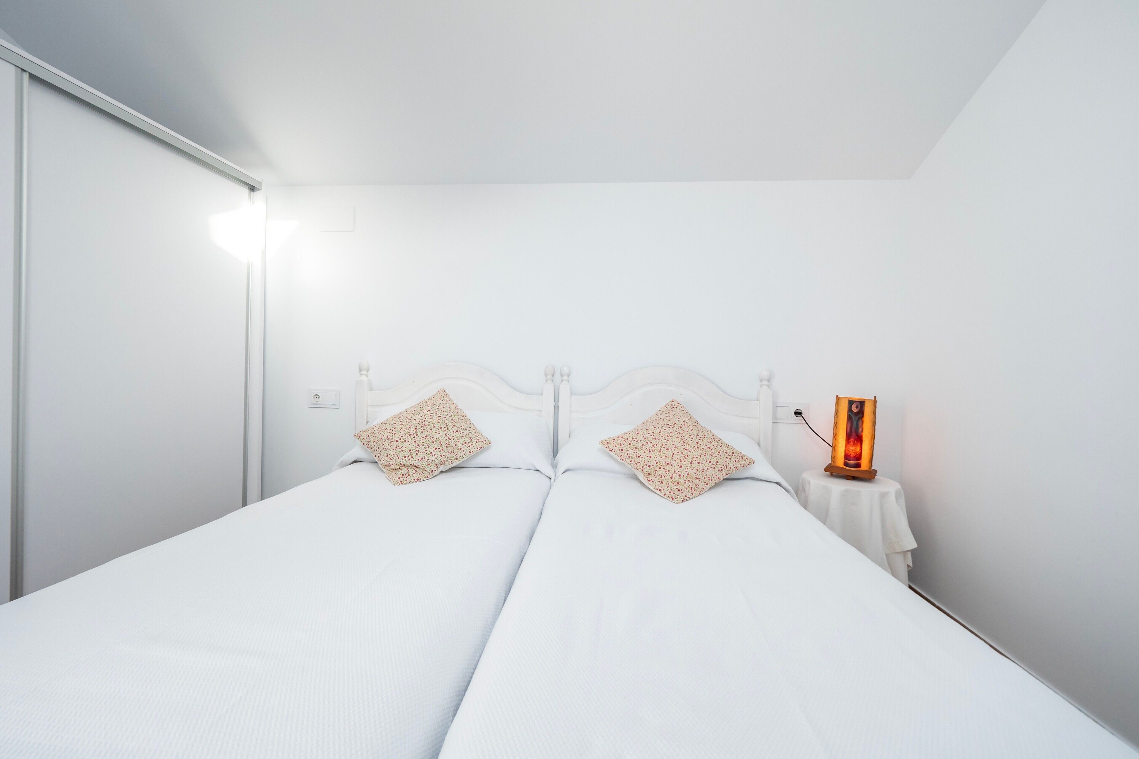 A double bed with a white headboard, two decorative pillows, a bedside table with a lamp, and a wall-mounted light.