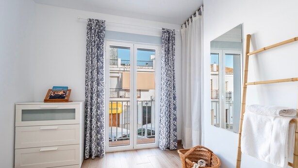 Quarto com cômoda branca, cesta de vime, porta-toalhas de bambu e uma porta de vidro deslizante com cortinas estampadas.