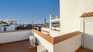 Terraço no telhado com parede branca, chão de azulejos marrons e vista para uma torre distante.