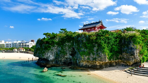 A coastal area with a traditional building on a cliff, a beach, and clear turquoise water. - Mr.KINJO in Kokusaidori (Naha)