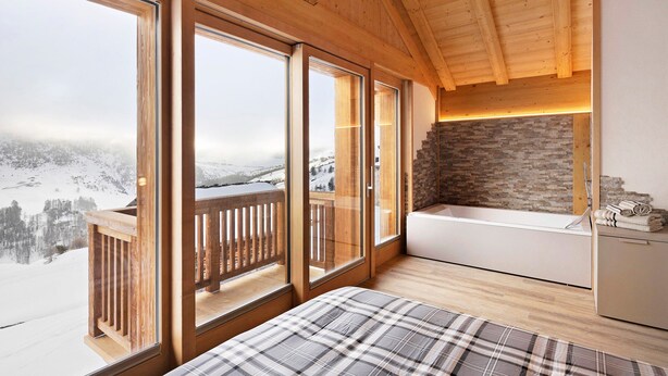 Una stanza con ampie finestre, soffitto in legno, parete in pietra, terrazza in legno e vasca da bagno bianca.
