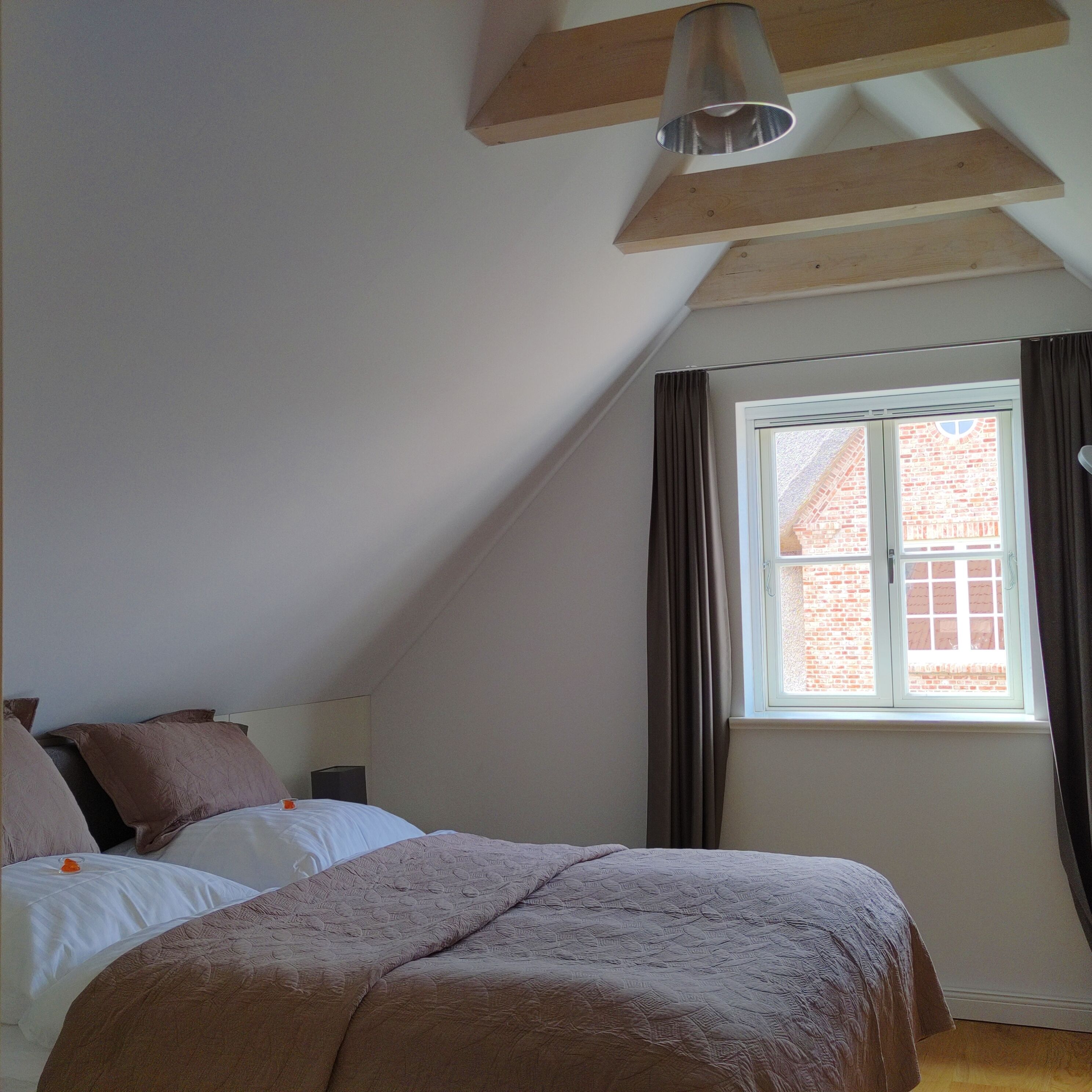 A bedroom with a sloped ceiling, a window with curtains, a bed with a brown blanket, and a bedside table.
