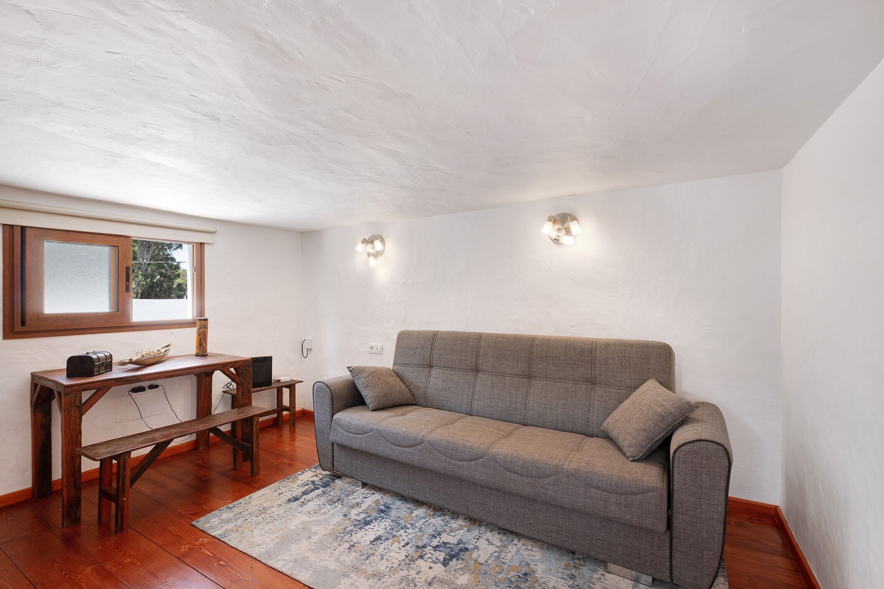 A room with a grey sofa, wooden table, bench, and a window with blinds.