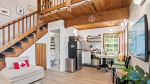 A modern kitchen with a dining area, wooden staircase, and a cozy living room with a sofa and a Canadian flag pillow. - Steps to Terrace Beach - Cozy Pet-Friendly Loft ⎮ Blue Heron (Ucluelet)