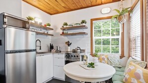 A modern kitchen with white cabinets, a stainless steel refrigerator, and a dining area with a round table and green sofa. - Steps to Terrace Beach - Cozy Pet-Friendly Loft ⎮ Blue Heron (Ucluelet)