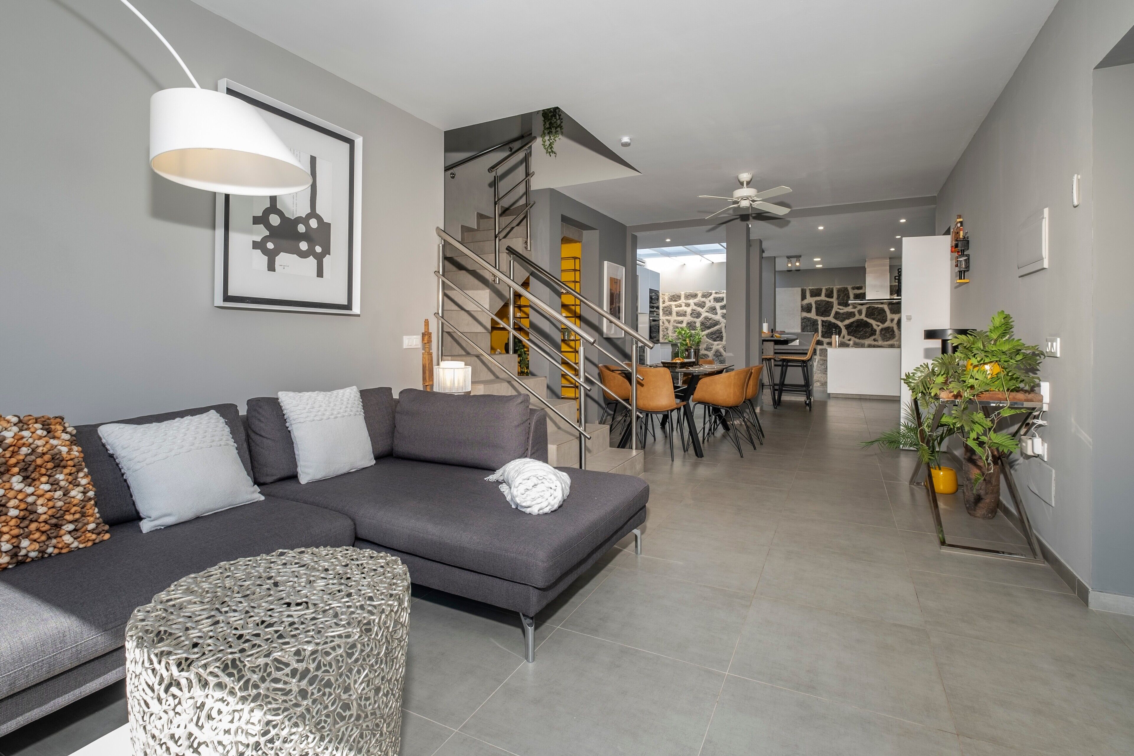 Een moderne woonkamer met een grijze hoekbank, een rond geweven pouf, een trap met een metalen leuning, en een eethoek met stoelen en een tafel.
