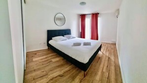 A bedroom with a bed, wooden flooring, a round mirror, and red curtains. - Charming renovated T2 Nogent-sur-Marne: Mobility lease (Nogent-sur-Marne)
