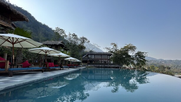 Una piscina a sfioro tranquilla con lettini prendisole in legno e ombrelloni, circondata da vegetazione rigogliosa e con le montagne sullo sfondo.