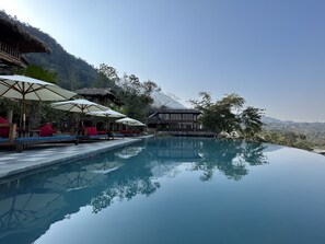 A serene infinity pool with wooden lounge chairs and umbrellas, surrounded by lush greenery and a mountain backdrop.