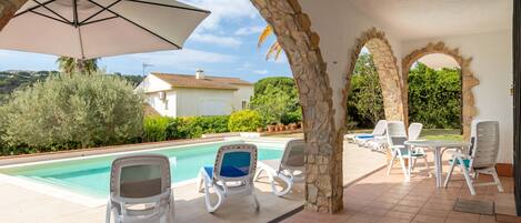 A poolside area with stone arches, lounge chairs, and a view of a house and greenery.