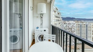 Een balkon met een wasmachine, een witte stoel en een raam met uitzicht op hoge gebouwen.