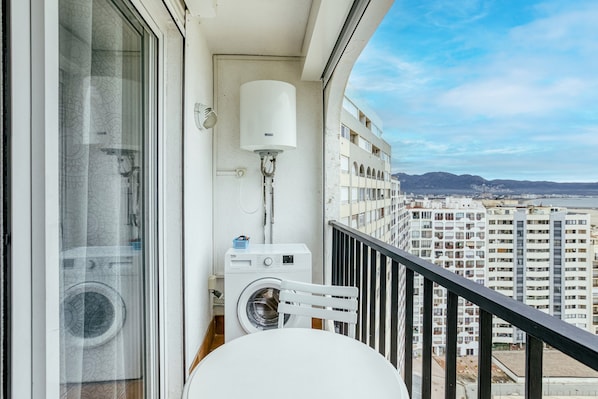A balcony with a washing machine, a white chair, and a window with a view of high-rise buildings.