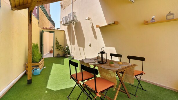 A patio with artificial grass, a wooden table, foldable chairs, a lantern, and a small shelf with decorative items.