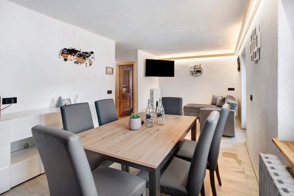 Un comedor moderno con una mesa de madera, sillas tapizadas en gris, una pequeña planta en maceta y un televisor de pantalla plana montado en la pared.