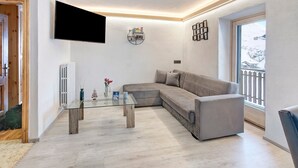 A modern living room with a sectional sofa, a glass coffee table, a mounted TV, and a window with a view of a snowy landscape. - Apartment 'Presot' with Mountain View, Shared Garden & Wi-Fi (Livigno)