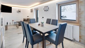 A modern dining room with a wooden table and chairs, a mounted TV, and a window with a snowy view. - Apartment 'Presot' with Mountain View, Shared Garden & Wi-Fi (Livigno)