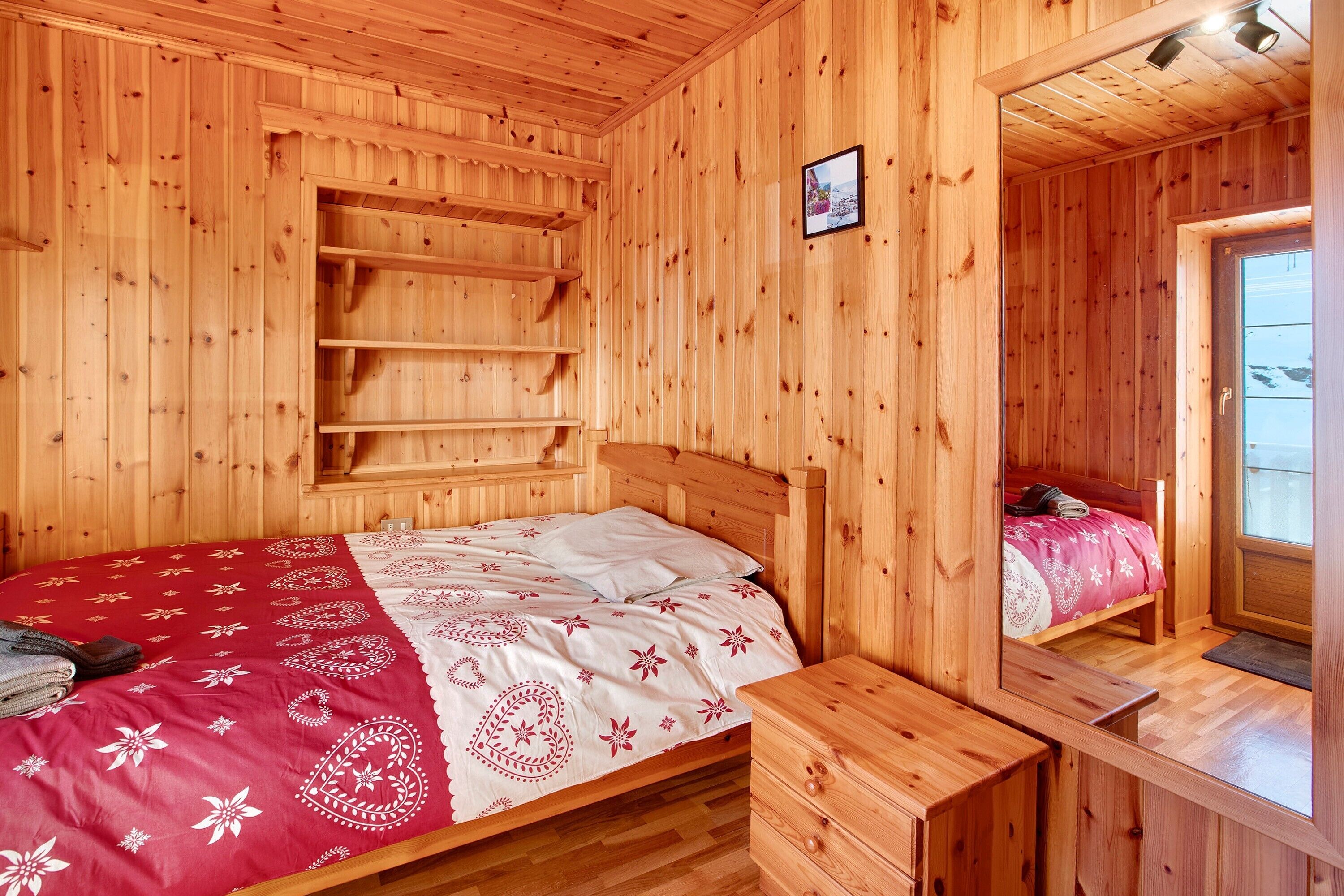 A wooden cabin bedroom with a bed, a dresser, a shelf unit, and a mirror.