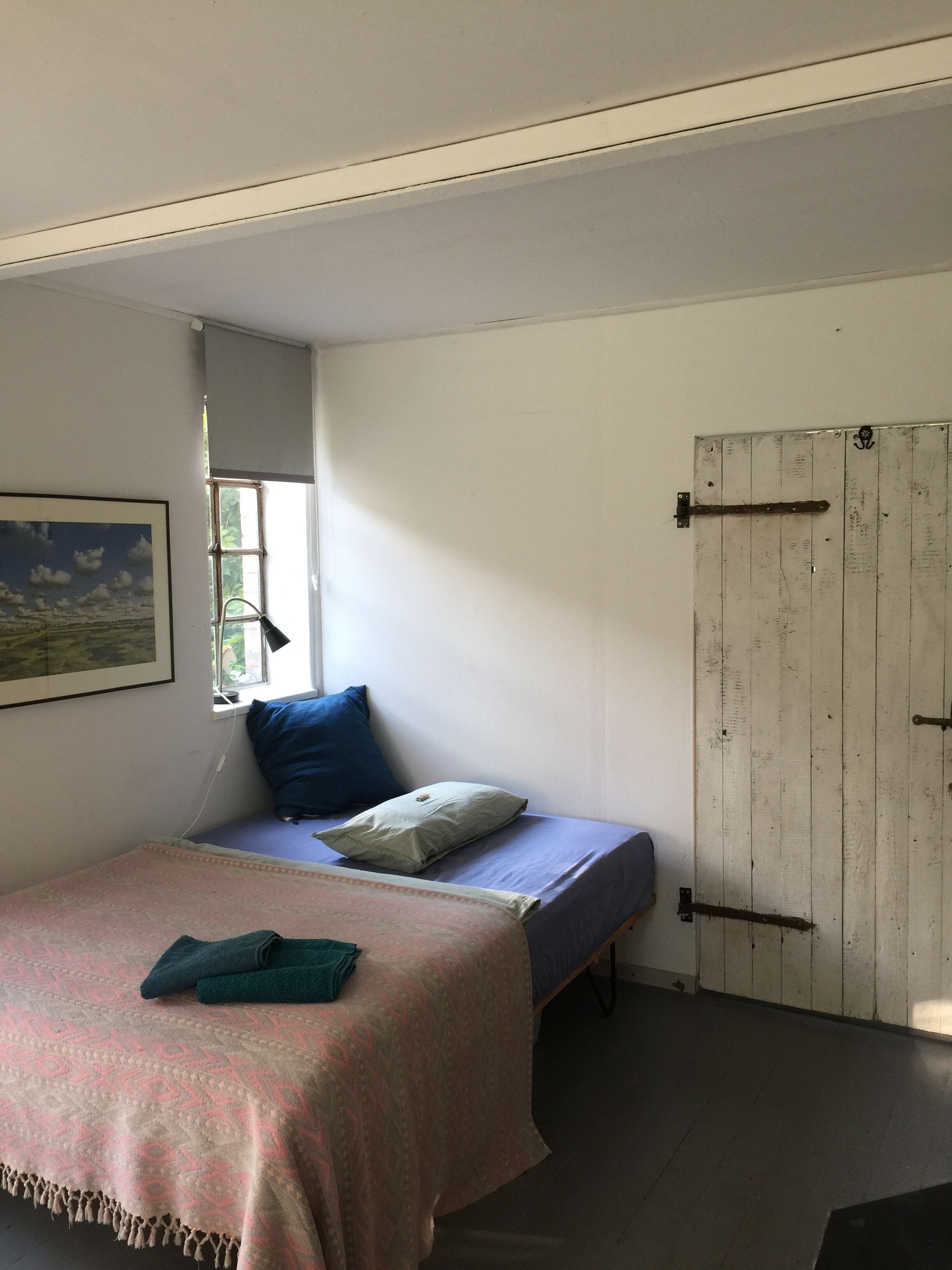 Ein Einzelbett mit einem gemusterten Bettüberwurf, einem blauen Kopfkissen und einem grünen Handtuch. Ein Fenster bietet Blick auf Grünflächen.