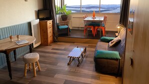 A cozy living area with a sofa, a coffee table, a TV, and a dining table with chairs. There is a view of the outdoors through large windows.