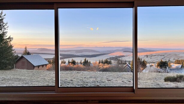 A view through a window with a wooden frame, displaying a landscape with rolling hills, sparse vegetation, and a small building with a sloped roof.