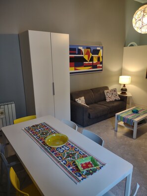A dining area with a white table, chairs, and a colorful table runner. There is a gray sofa with patterned cushions and a lamp on a side table. A colorful abstract painting is on the wall.