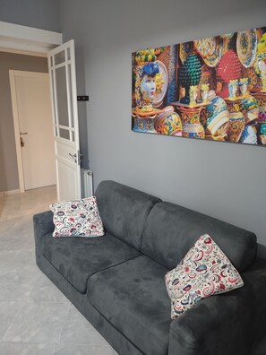 A grey sofa with two patterned cushions, a colorful abstract painting on the wall, and a partially open white door in the background.