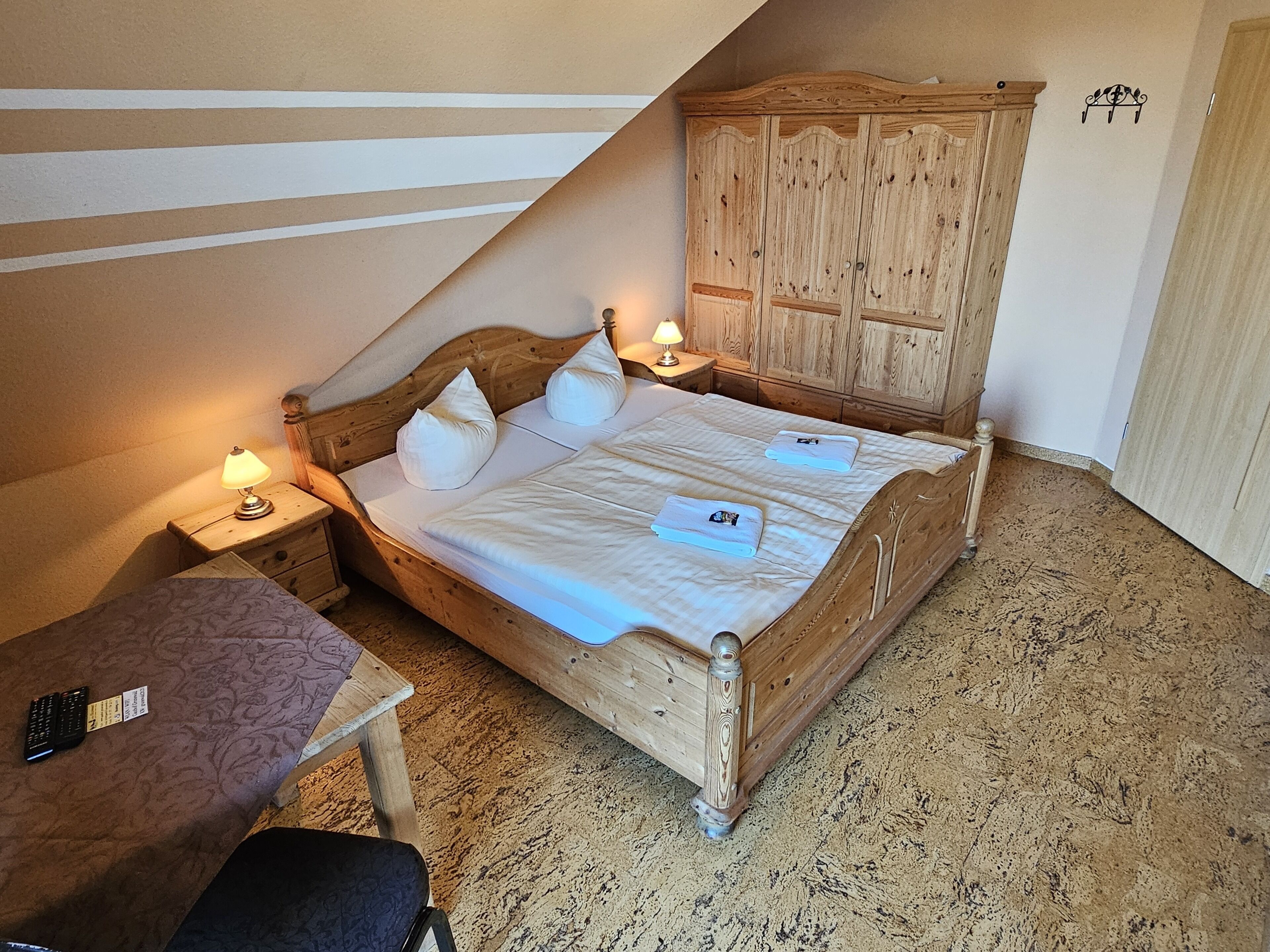 A wooden bed with two pillows and a nightstand with a lamp, in a room with a sloped ceiling and a wooden wardrobe.