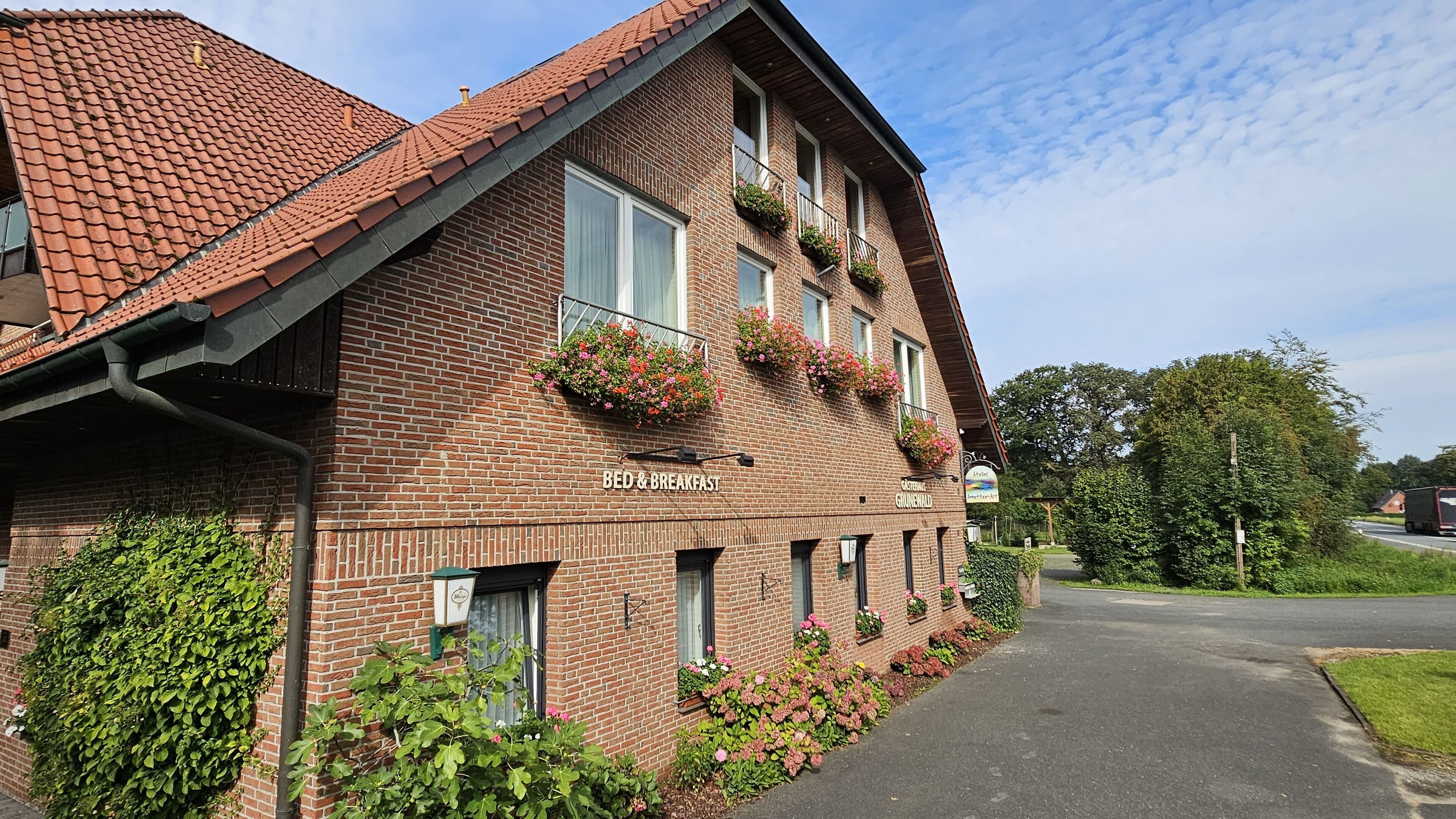 A brick building with a sign reading "BED & BREAKFAST" and "Grünewald." It has multiple windows with flower boxes and a paved driveway.