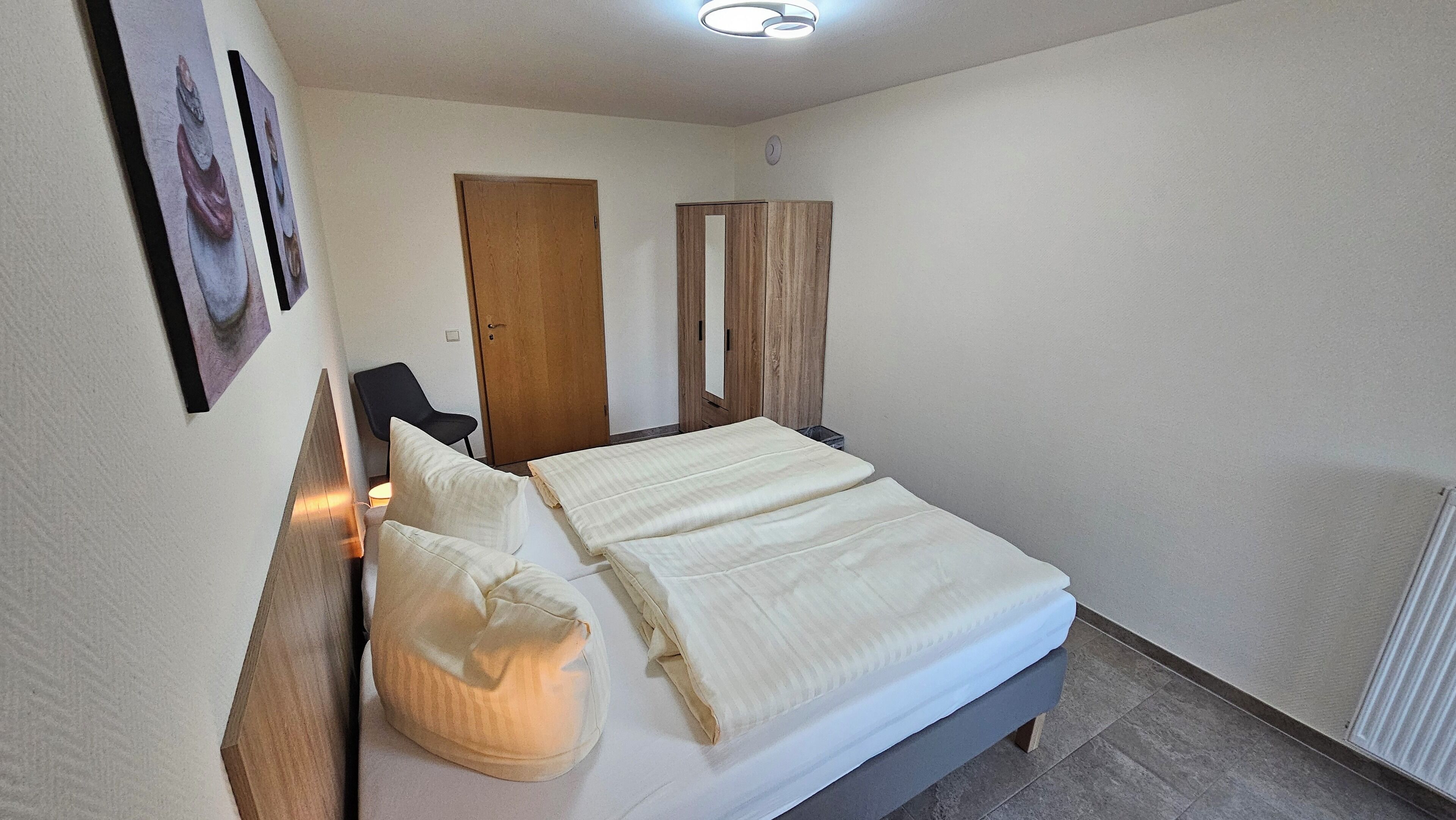 Una cama bien hecha con almohadas y una manta, un armario de madera, una silla y un aplique de pared.