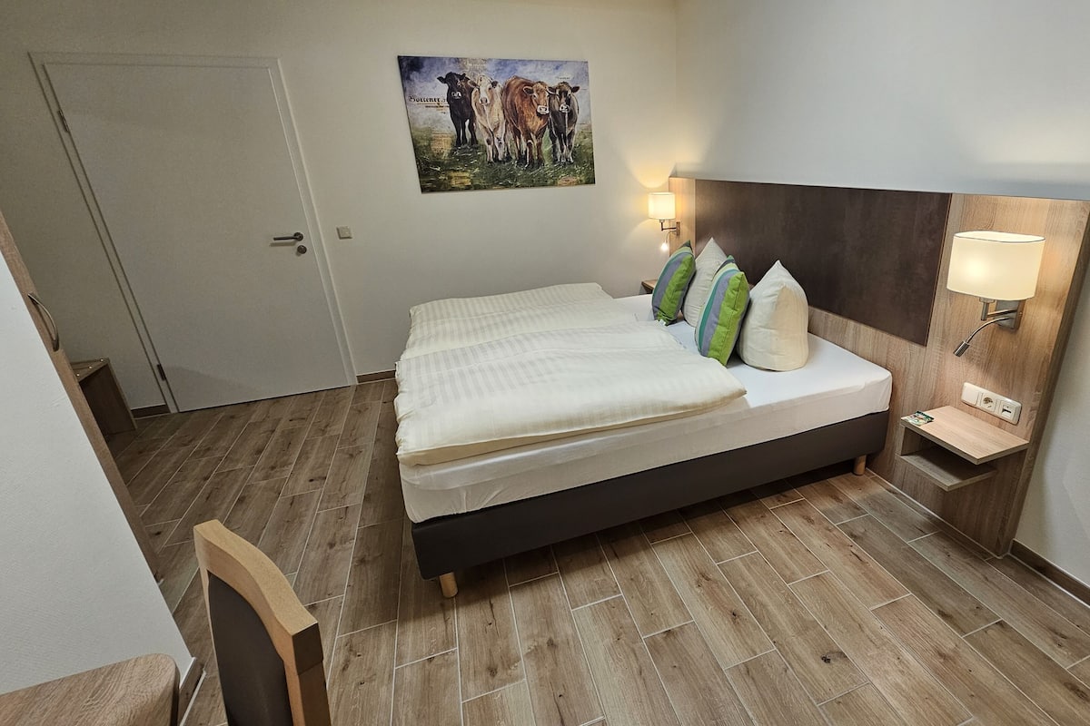 Uma cama bem arrumada com cabeceira, uma mesa de cabeceira com abajur e uma obra de arte na parede com tema de vacas.