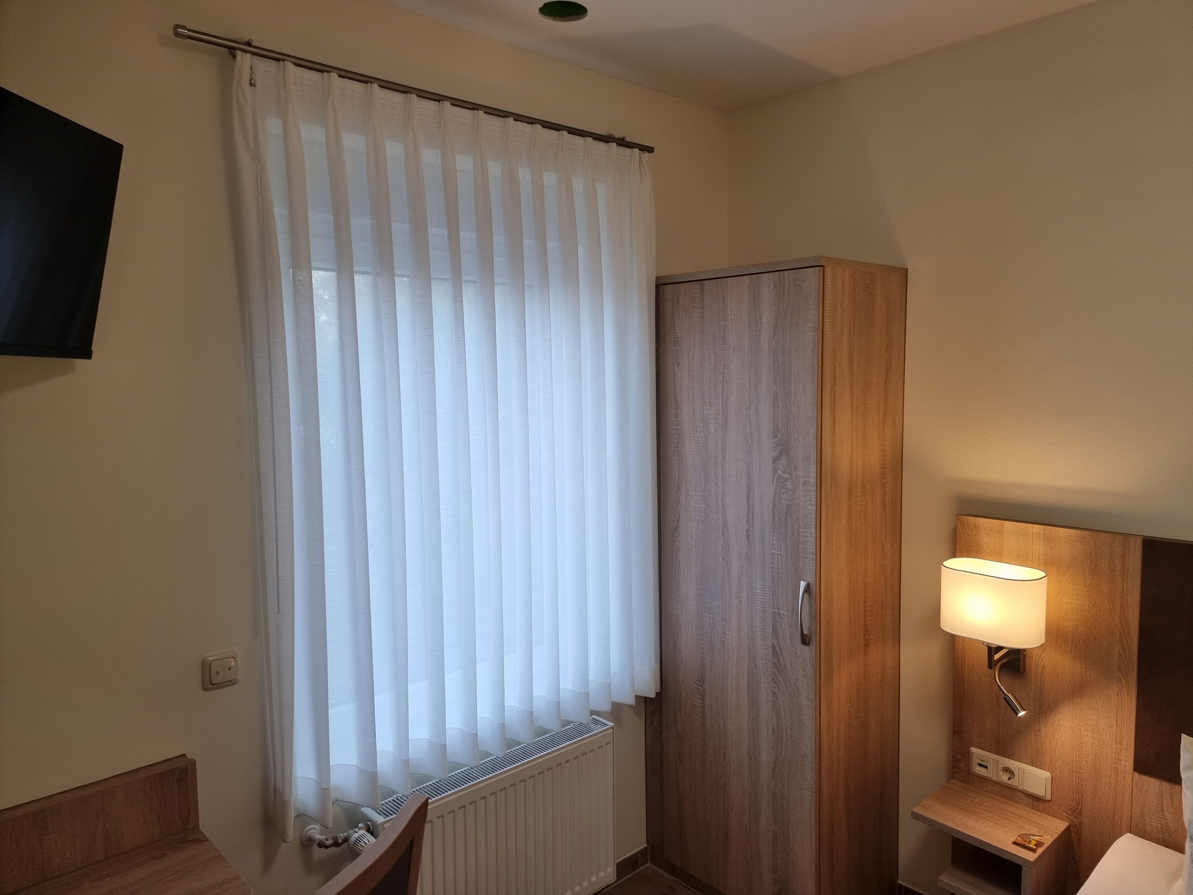 A hotel room with a wooden wardrobe, a radiator, a bedside table with a lamp, and a TV mounted on the wall.